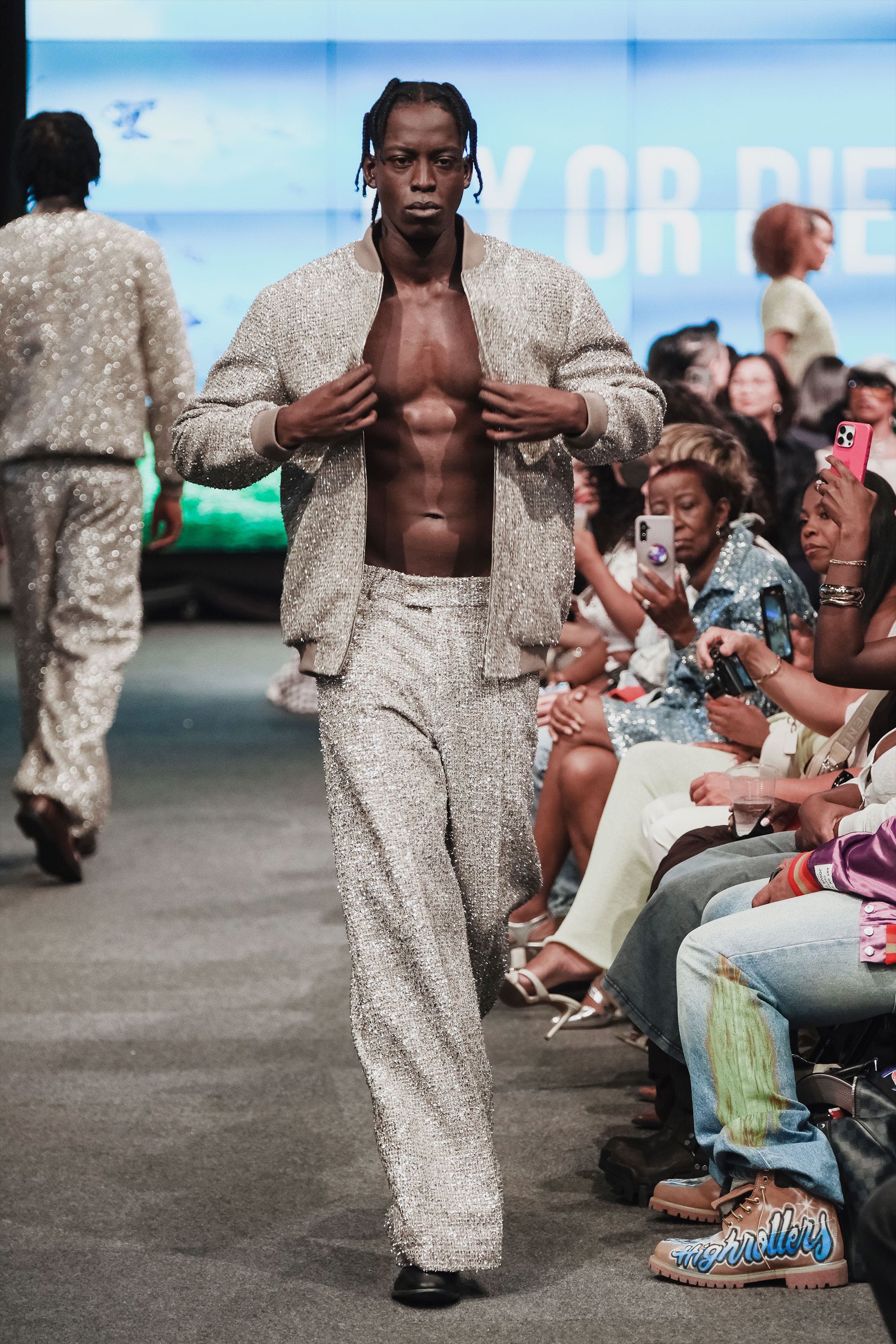 Model walking on a runway wearing a textured metallic tweed outfit with an audience watching.