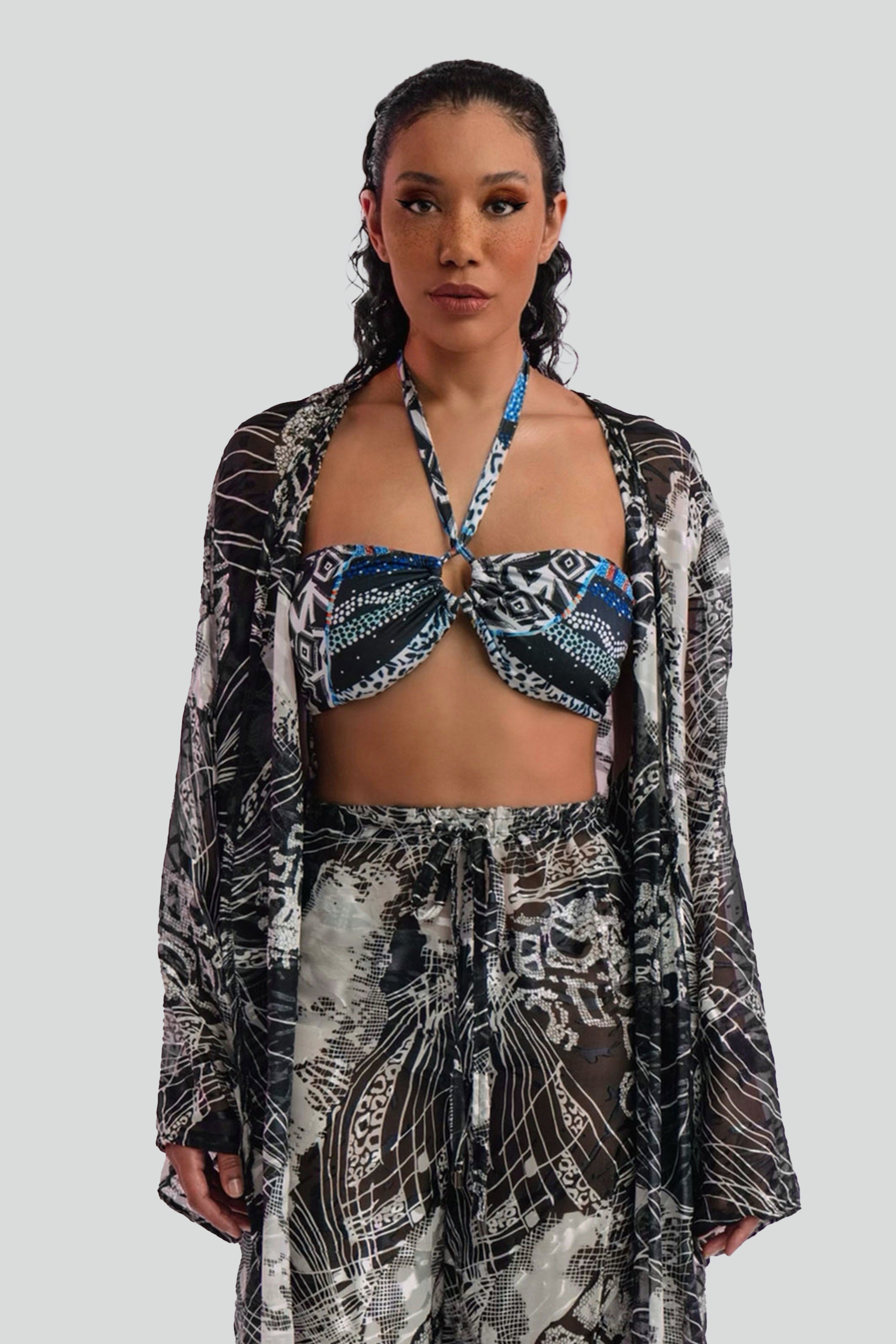 Woman wearing a long, black and white patterned cover up top over a matching bikini and pants set on a plain background