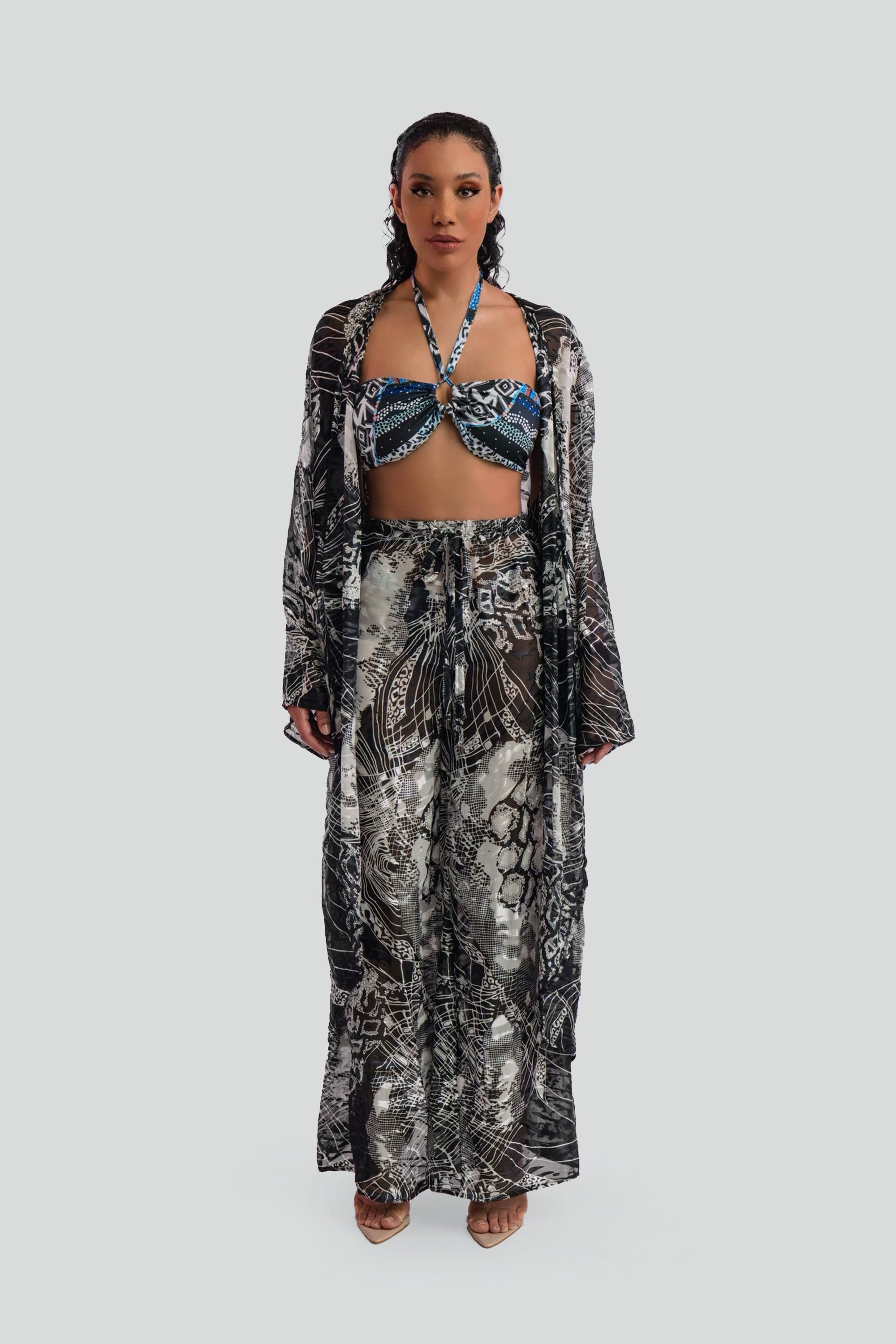Woman wearing a long, black and white patterned cover up top over a matching bikini and pants set on a plain background