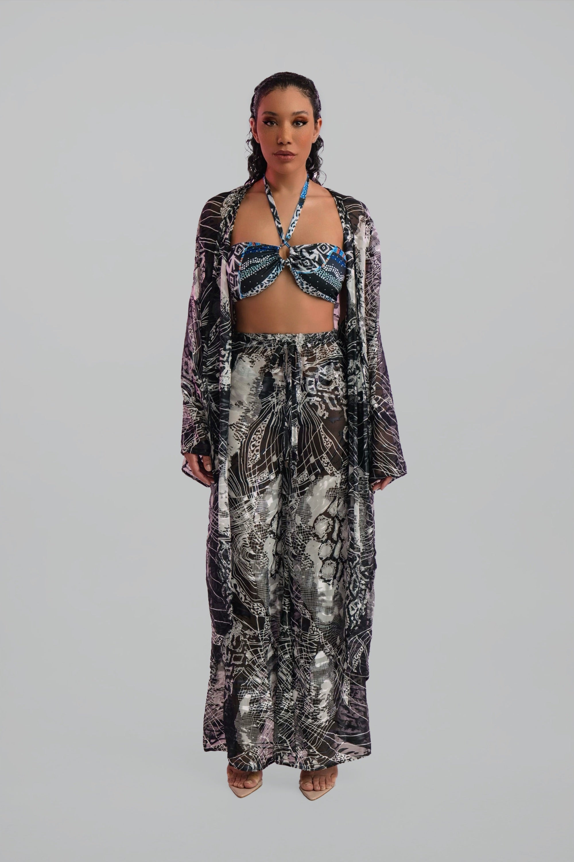 Woman wearing a long, black and white patterned cover up top over a matching bikini and pants set on a plain background