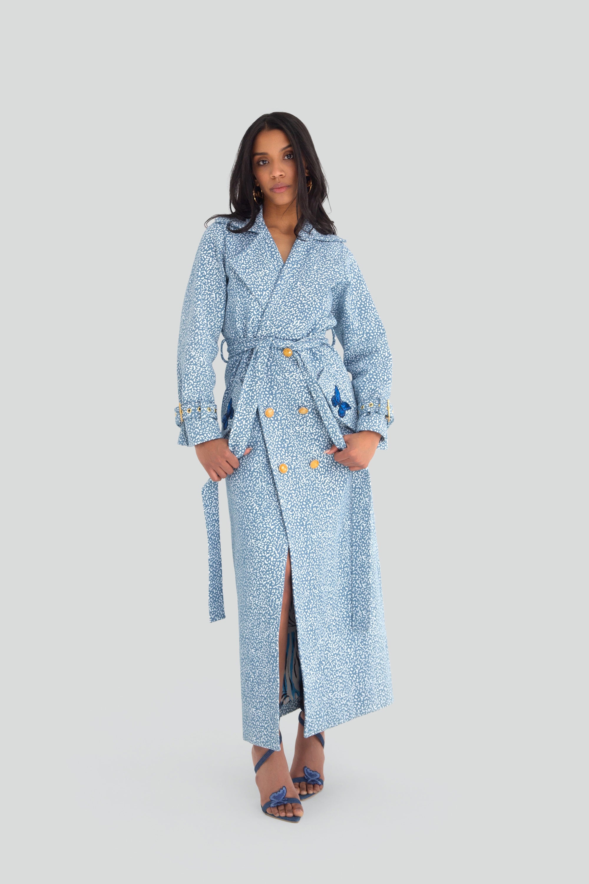 Woman wearing a blue patterned trench coat with gold buttons and butterfly details on a plain background