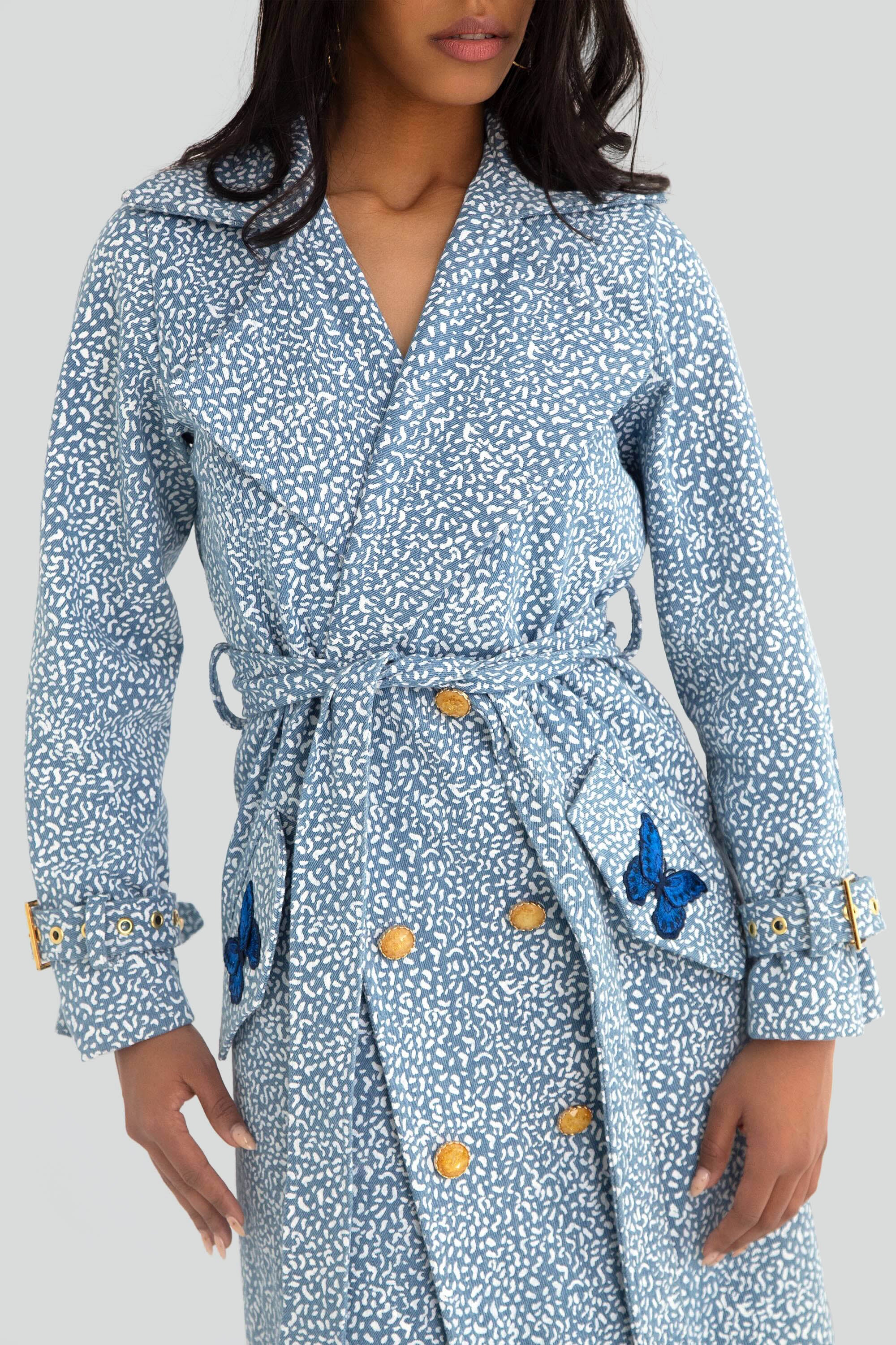 Woman wearing a blue patterned trench coat with gold buttons and butterfly details on a plain background