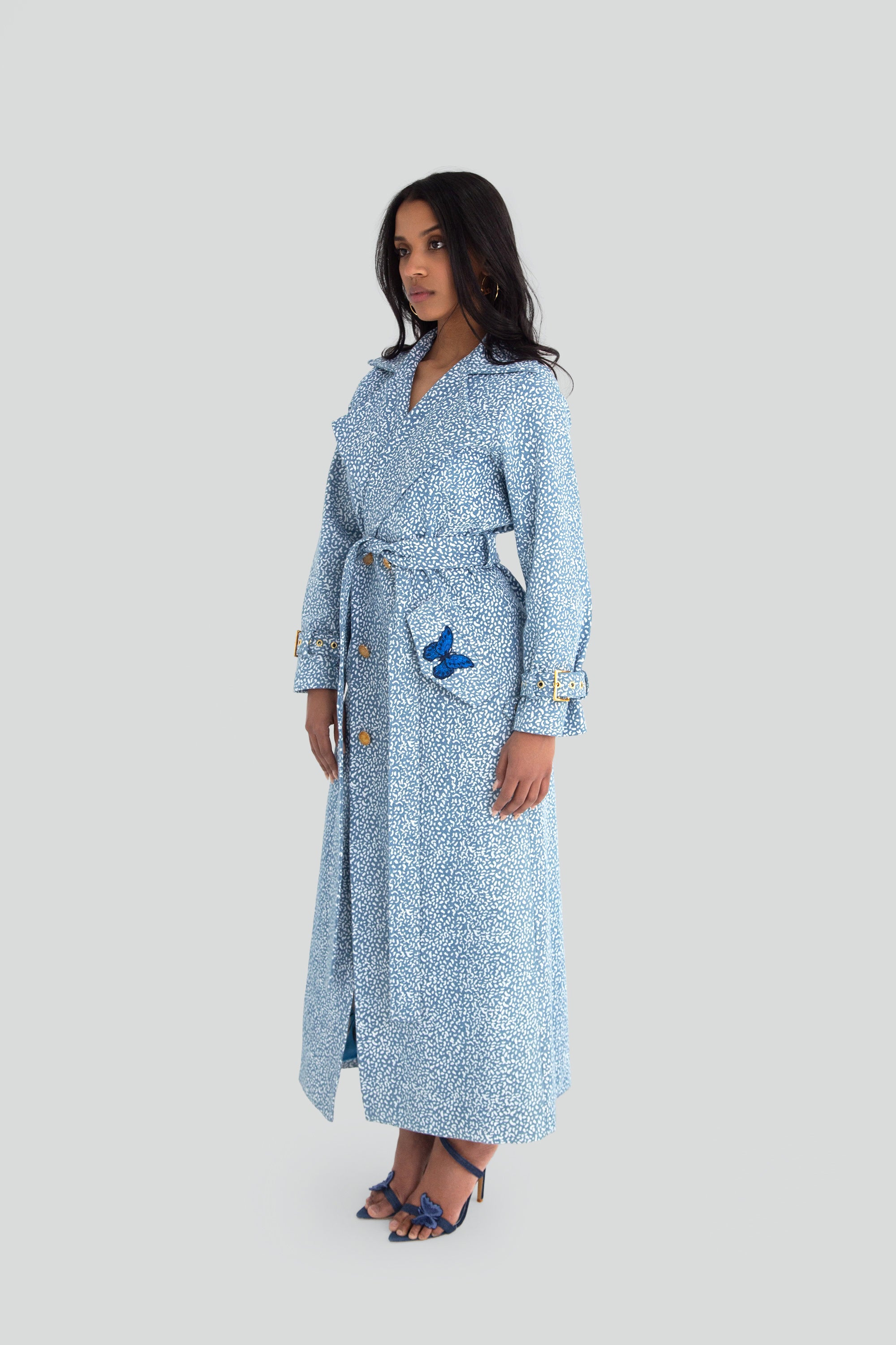 Woman wearing a blue patterned trench coat with gold buttons and butterfly details on a plain background