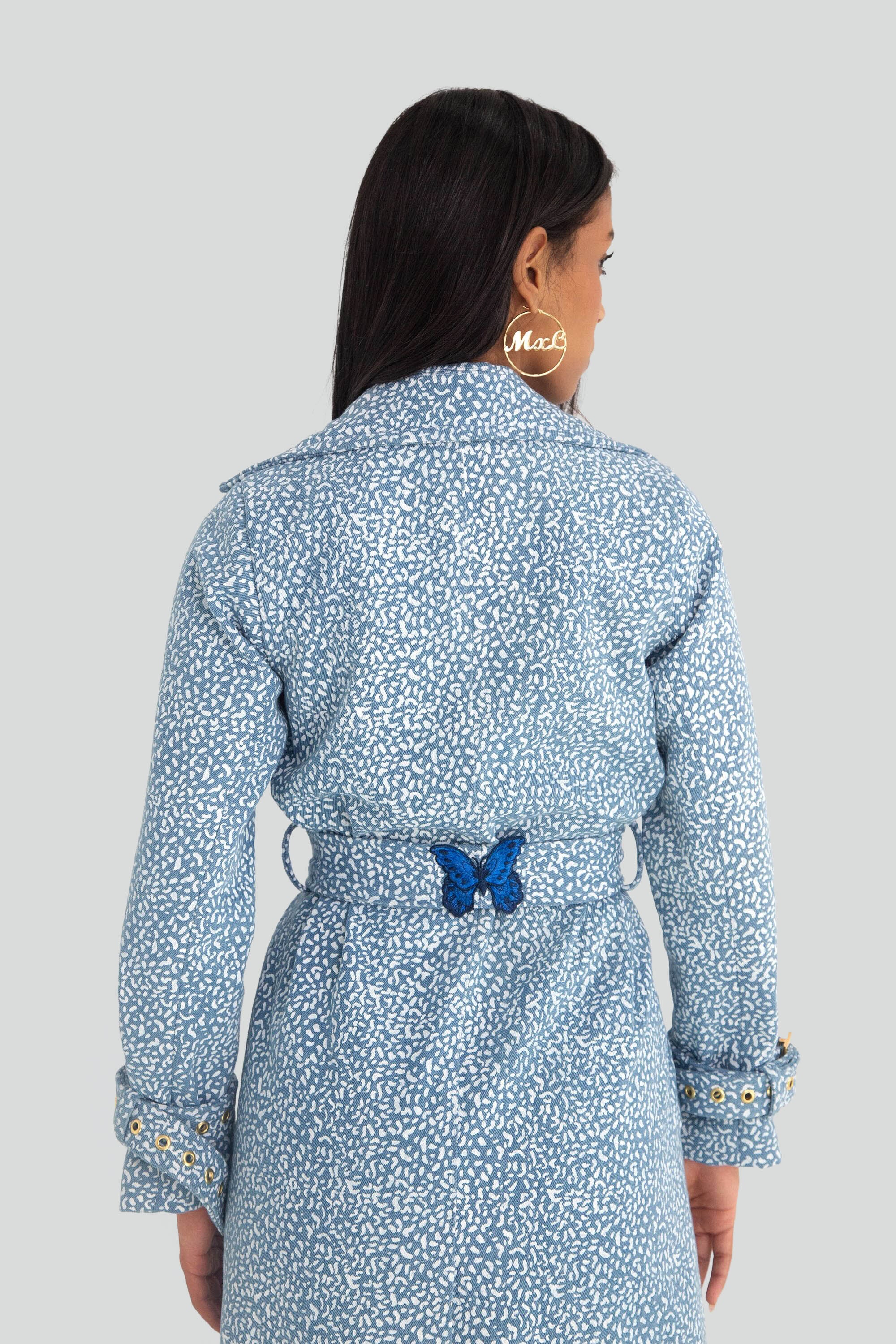 Woman wearing a blue patterned trench coat with gold buttons and butterfly details on a plain background