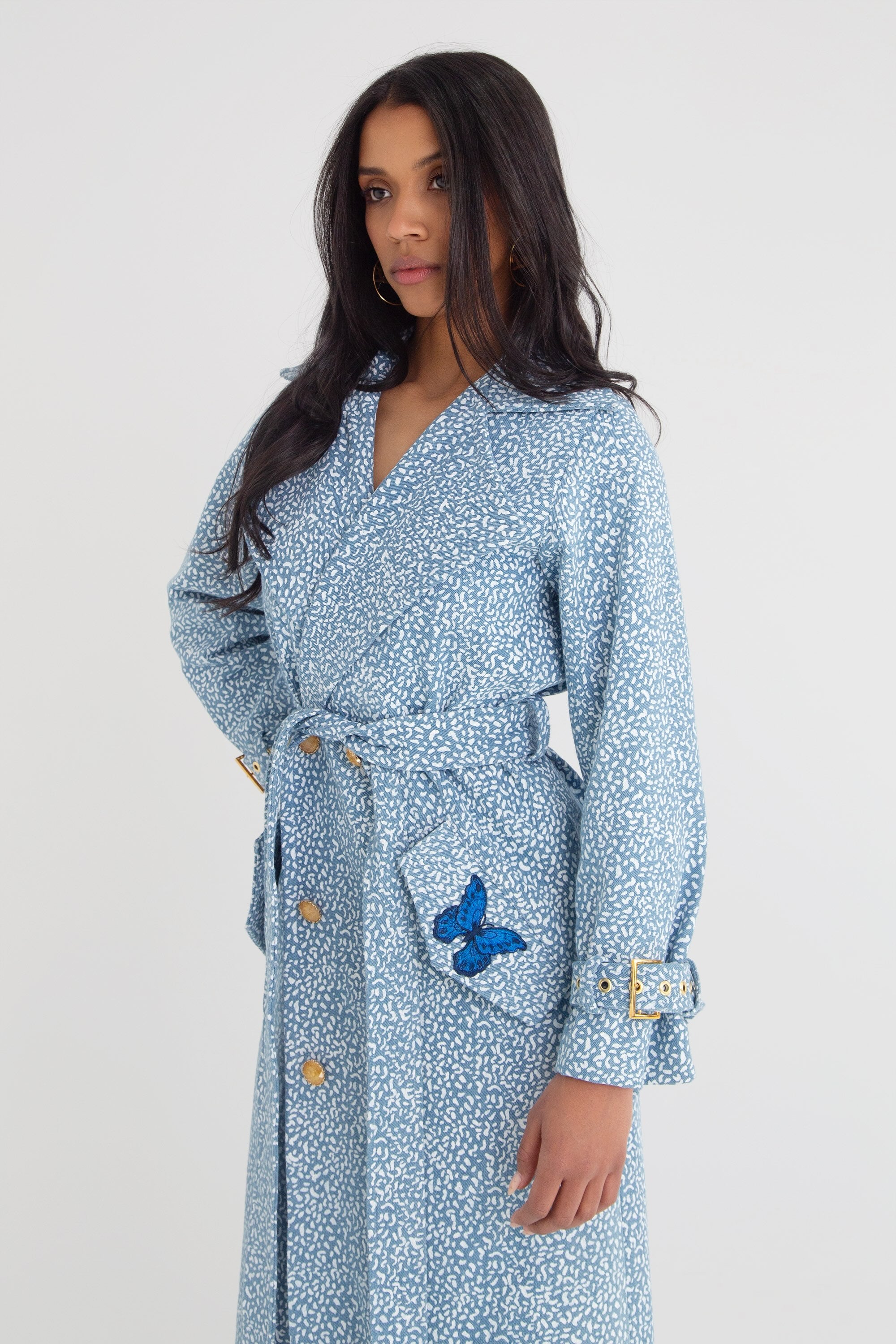 Woman wearing a blue patterned trench coat with gold buttons and butterfly details on a plain background
