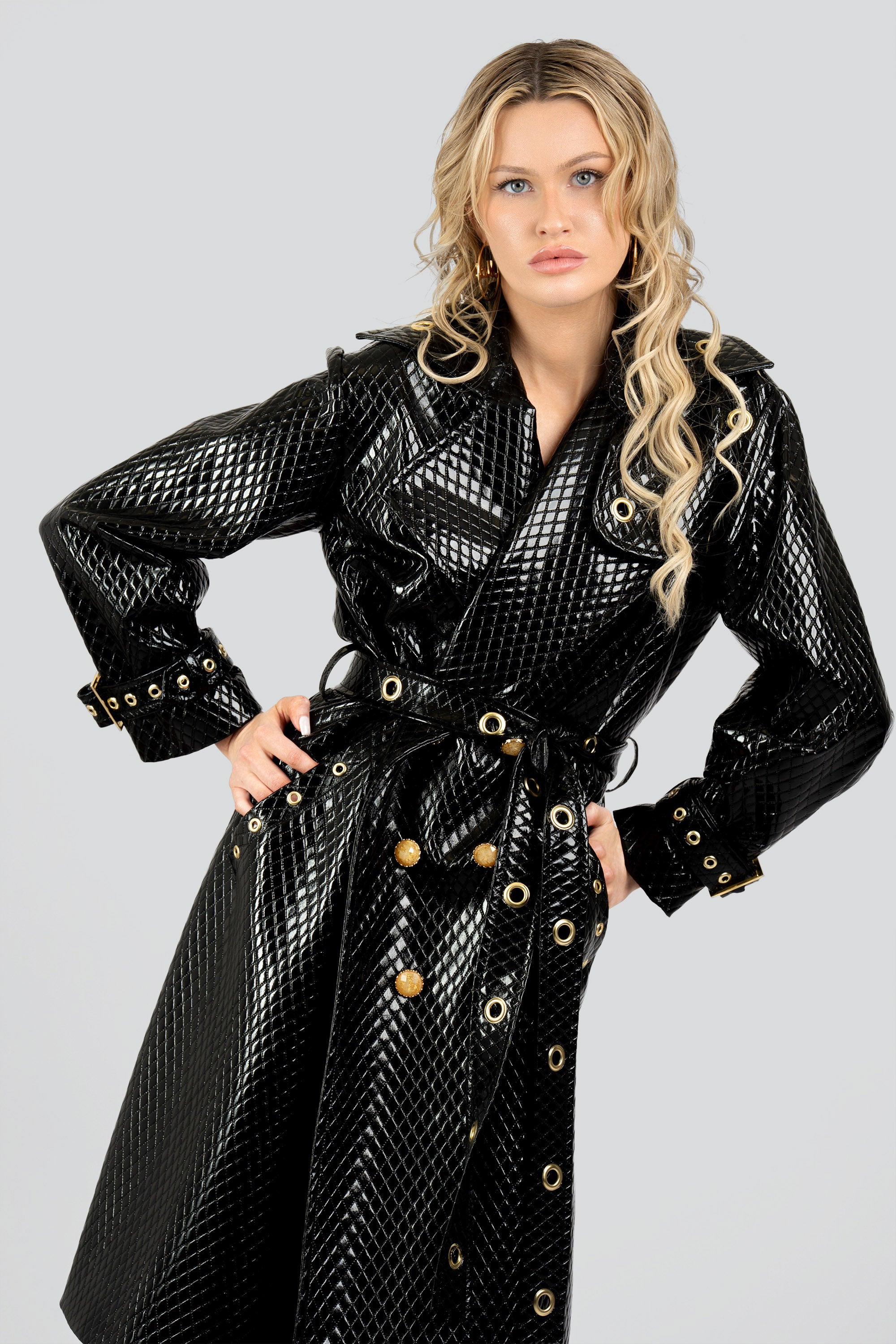 Woman wearing a quilted black trench coat with a belt and gold accents on a white background
