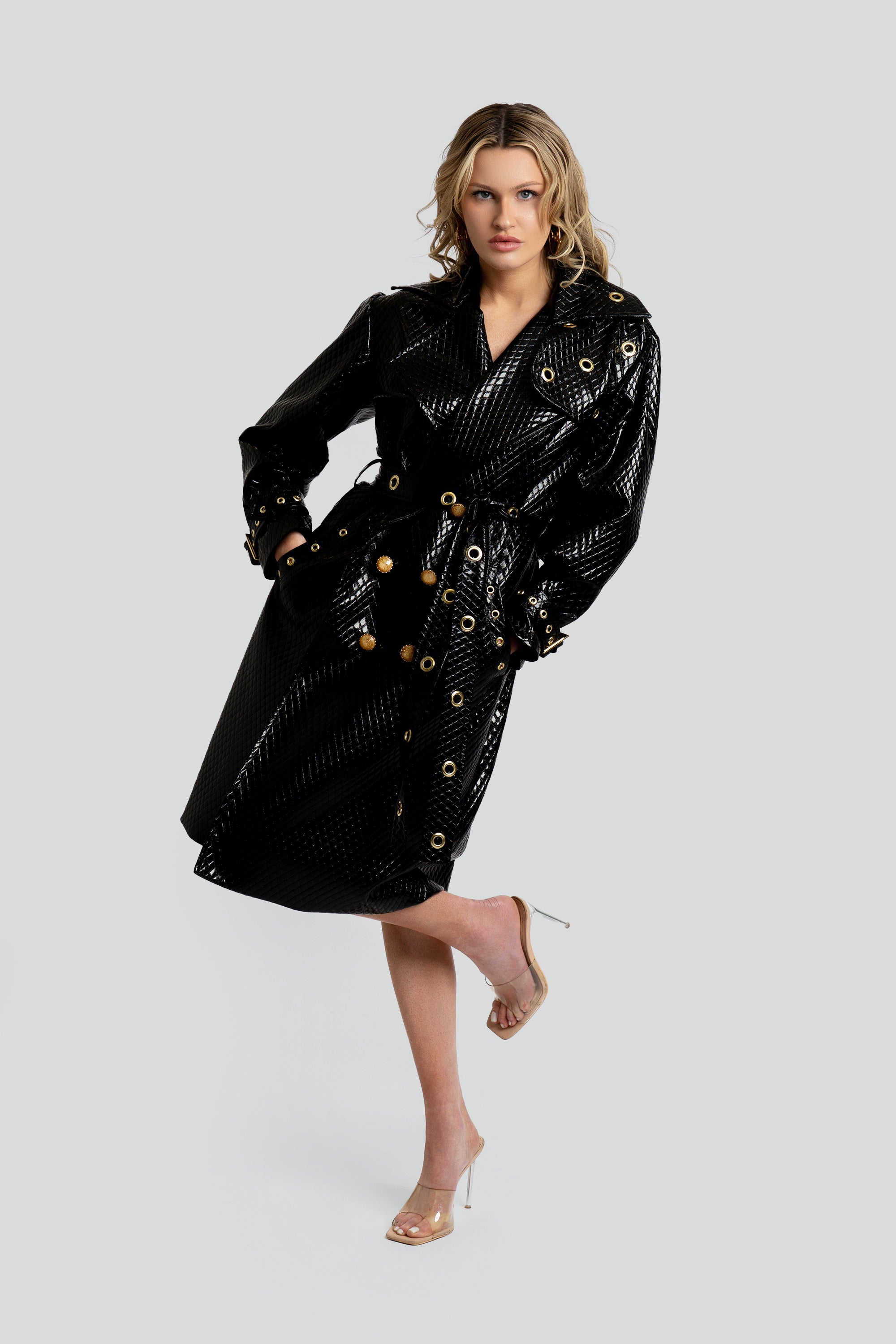 Woman wearing a quilted black trench coat with a belt and gold accents on a white background