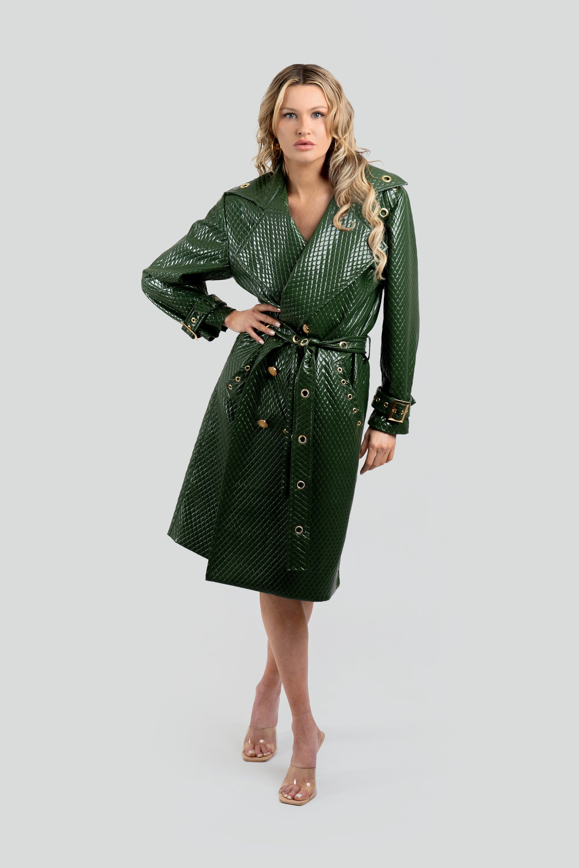 Woman wearing a quilted green trench coat with a belt and gold accents on a white background