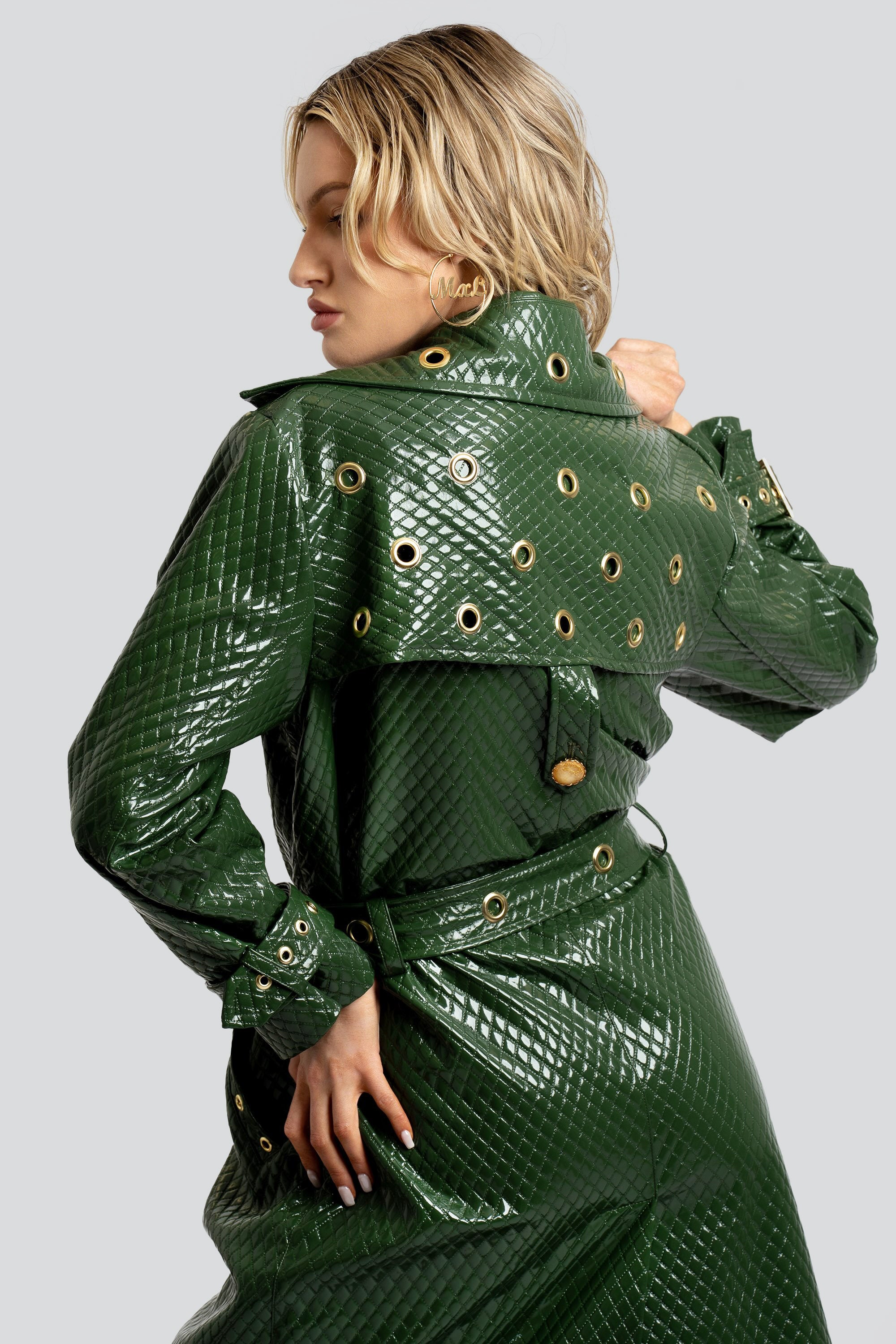 Woman wearing a quilted green trench coat with a belt and gold accents on a white background
