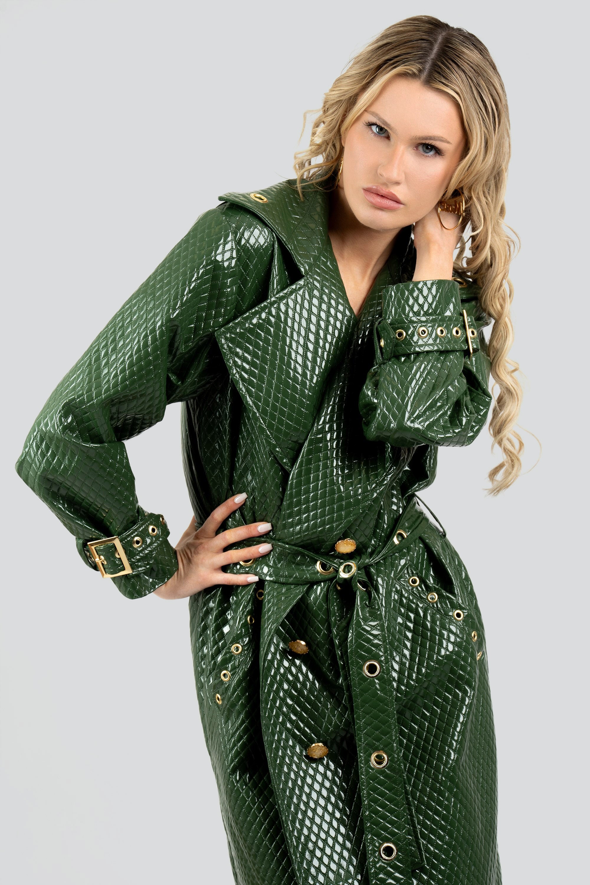 Woman wearing a quilted green trench coat with a belt and gold accents on a white background