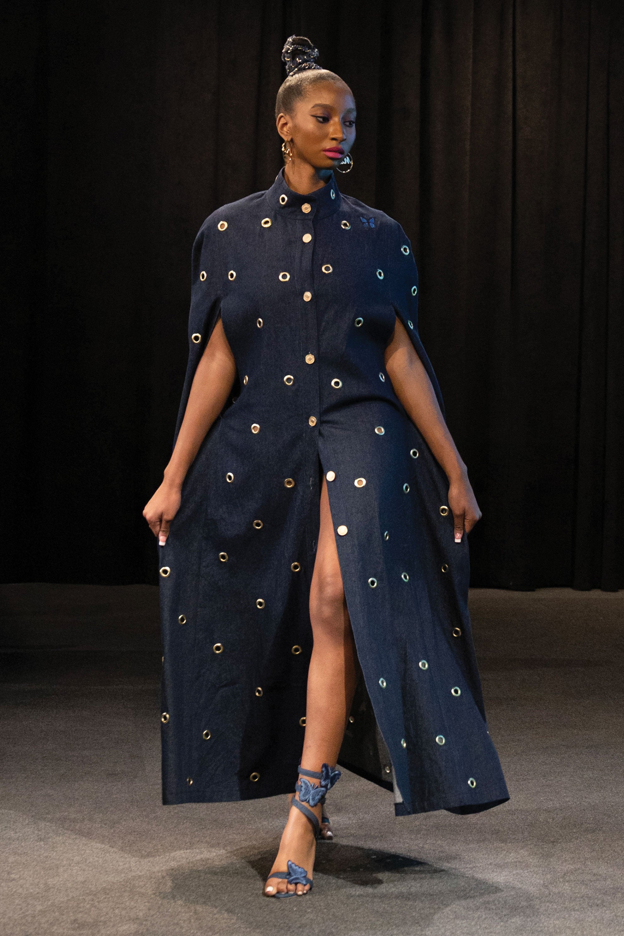 Model wearing a dark blue dress with gold embellishments against a black background